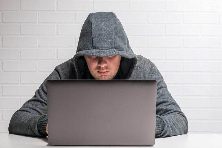 A man in a hoodie is using a laptop to blog anonymously.