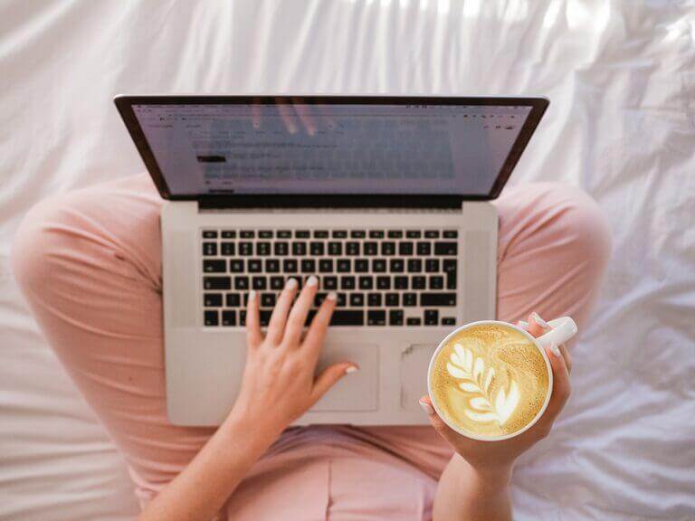 A woman is sitting on a bed with a laptop and a cup of coffee, pondering how many blog posts to write per month.